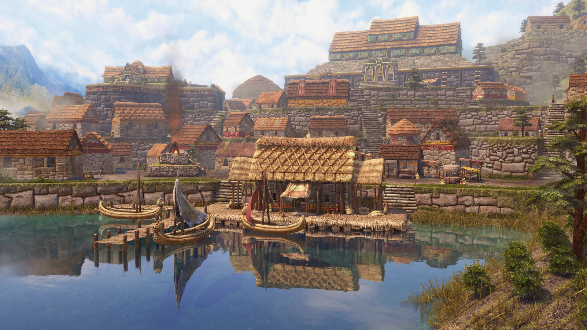A side view of a civilization from Age of Empires III: Definitive Edition. A large body of water is at the front of the image with a dock and two fishing boats docked. Beyond the dock are several large stone buildings with brown thatched roofs. The ground has multiple layers that keep getting higher. With each layer are more buildings, reaching up toward the largest building in the distance. Mountains and white clouds are in the distance.